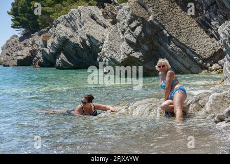 Zwei Frauen posieren in klarem, transparentem Meerwasser auf den hellen Steinen eines flachen Strandes von Lucice in der Nähe von Petrovac, Montenegro. Stockfoto