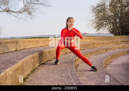 Junge Fitness-Frau im roten Sport trägt dabei Seitensprung, Kniebeugen beim Training im Freien auf der Stadiontreppe. Stretching nach dem Lauftraining. Aktiv Stockfoto