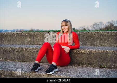 Porträt einer lächelnden, selbstbewussten Fitness-Frau in rotem Sportkleidung, die sich nach dem Training ausruhte oder bei Sonnenuntergang im Freien lief. Joggen im Stadion. Aktiv Stockfoto