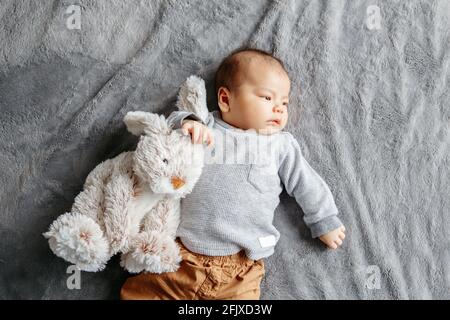 Zwei Monate neugeborener asiatischer Junge auf dem Bett liegend mit Kaninchen-Komfortspielzeug Stockfoto