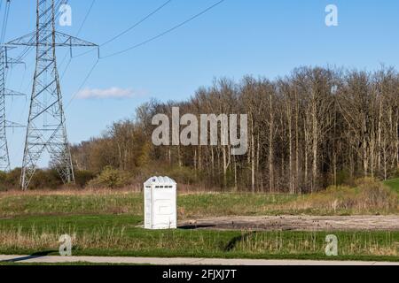 Weiße tragbare Toilette am Ende eines Parkplatzes und neben einem Feld und hydroelektrischen Türmen. Wald im Hintergrund. Stockfoto