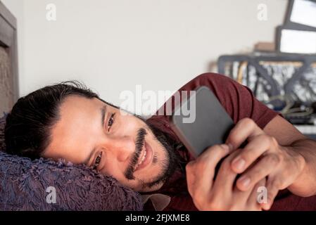 Junger lateinischer Mann, der lächelte und das Telefon auf dem Bett benutzte Zuhause bei Tageslicht Stockfoto