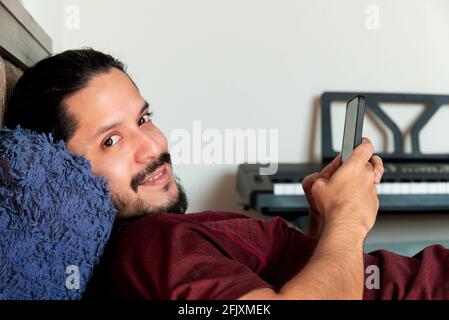 Junger lateinischer Mann, der lächelte und das Telefon auf dem Bett benutzte Zuhause bei Tageslicht Stockfoto