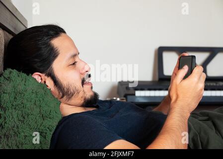 Junger lateinischer Mann, der lächelte und das Telefon auf dem Bett benutzte Zuhause bei Tageslicht Stockfoto