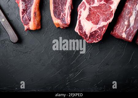 Auswahl an rohen Rindersteaks zum Grillieren mit Gewürz- und Utensilien-Set, Tomahawk, T-Bone, Clubsteak, Rib-Eye- und Filet-Schnitten, auf schwarzem ston Stockfoto
