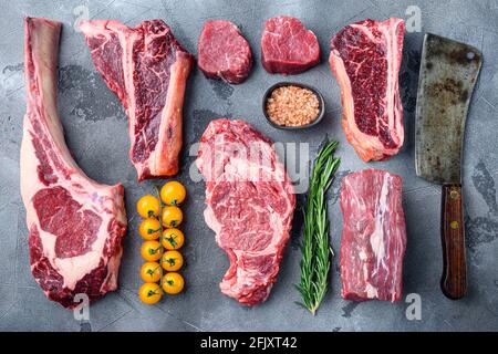 Auswahl an rohen Rindersteaks zum Grileln mit Gewürz- und Utensilien-Set, Tomahawk, T-Bone, Clubsteak, Rib-Eye- und Filet-Schnitten, auf grauem Stein Stockfoto