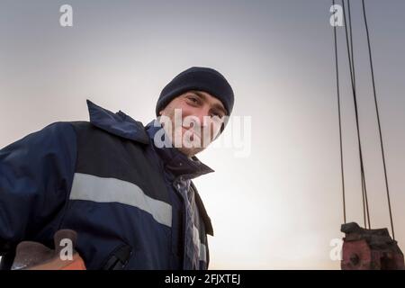 Porträt eines Arbeiters in einer blauen Jacke mit einem Haube am Haken eines LKW-Krans Stockfoto