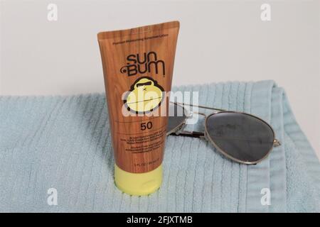 Sun Bum Sunscreen Lotion auf einem blauen Handtuch neben einer Sonnenbrille auf weißem Hintergrund. Stockfoto