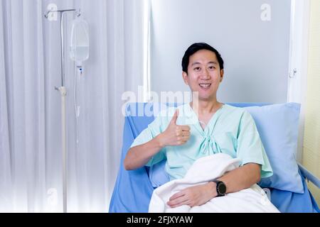 Porträt eines lächelnden asiatischen jungen männlichen Patienten sitzen auf Krankenhausbett Zimmer zeigt Daumen nach oben Geste und Zeichen zu Kamera Stockfoto