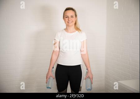 Glückliche blonde Frau macht Fitness-Übung mit Wasserflaschen - Basic Sport Workout als Training zu Hause Stockfoto