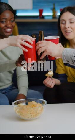 Multikulturelle Freunde geselligen sich, während sie spät in der Nacht auf dem Sofa im Wohnzimmer sitzen und Bier trinken und die Heimparty genießen. Eine Gruppe gemischter Leute, die Zeit miteinander verbringen Stockfoto