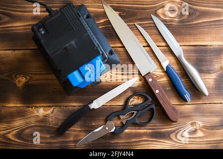 Schärfen eines Messers auf einem elektrischen Spitzer zu Hause. Anordnung von Messern und Scheren mit einer Schärfmaschine auf einem Holztisch. Draufsicht, flach liegend. Stockfoto