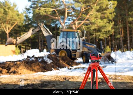 Eine geodätische Ebene steht vor einer Planierraupe zum Ausheben einer Grube auf einer Baustelle. Der Baggereimer gräbt ein Loch, um ein Haus zu bauen Stockfoto