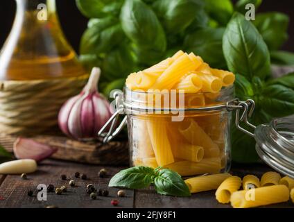 Rohe Penne Pasta im Glas mit Öl und Knoblauch, Basilikum-Pflanze auf Holztisch Hintergrund. Makro Stockfoto