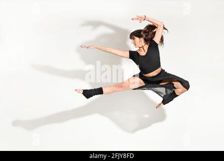 Agile junge Tänzerin, die einen Halbsprung vor der Tür vorführt Mit langen Haaren, die in einer vereinzelten Midair-Pose fliegen Weiß mit dramatischen Schatten und Copyac Stockfoto