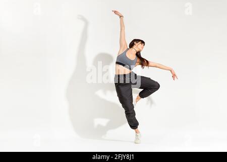 Anmutige junge Tänzerin in einem Hip-Hop-Outfit Eine klassische Tanzpose mit ausgestreckten Armen, die auf einem balancieren Bein mit künstlerischem Schatten auf einem Stockfoto