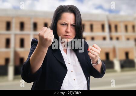 Verärgerte Geschäftsfrau im eleganten legeren formellen Anzug mit kämpferischen Fäusten Als aggressive Geste bei der Renovierung des Gebäudes im Hintergrund Stockfoto