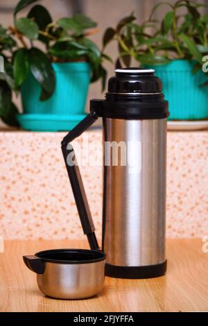 Thermoskanne mit einem Becher auf dem Tisch im Raum auf dem Hintergrund von Zimmerpflanzen. Selektiver Fokus Stockfoto