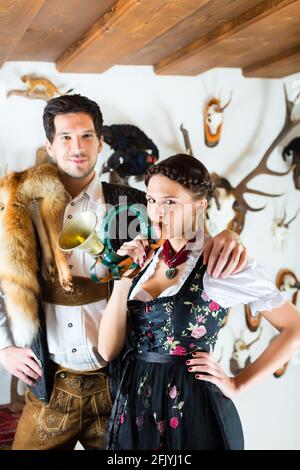 Junger Jäger mit seiner Frau und einer Trompete vor einer Wand mit verschiedenen Hörner, Geweihe und Trophäen in einer Almhütte Stockfoto