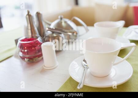 Tischeinstellung im Café. Becher mit Untertasse, Teedose Tee, Salz, Pfeffer, Zucker und Vase mit Rose auf dem Tisch. Frühstück oder Tee im Restaurant. Stockfoto