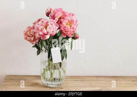 Blumenstilles Leben Szene. Rosa Pfingstrosen Blumen, Blumenstrauß in Glasvase auf Holztisch. Leerer Geschenkanhänger, Etikettenmockup. Weißer Wandhintergrund. Seelektiv Stockfoto
