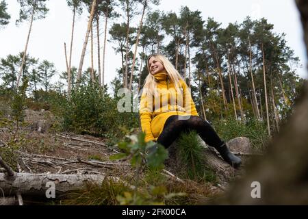 Blonde Frau im gelben Mantel sitzt im Herbstwaldes. Entspannen im Wald Stockfoto