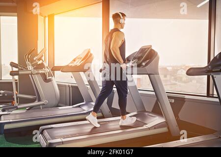 Konzentrierter, fitter Mann, der in Kopfhörern Musik hört, während er auf dem Laufband in einem modernen Fitnessstudio mit Panoramafenstern läuft. Stockfoto