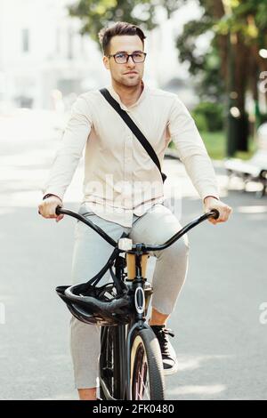 Moderner Büroangestellter, persönlicher Ökotransport in der Stadt und aktiver Lebensstil. Serious Millennial Attraktive männlich in Brille mit Tasche Fahrten auf dem Fahrrad in Stockfoto