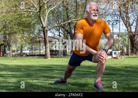 Ein älterer, selbstbewusster Mann trainiert im Park, dehnt sich die Beine und schaut nach vorne. Stockfoto