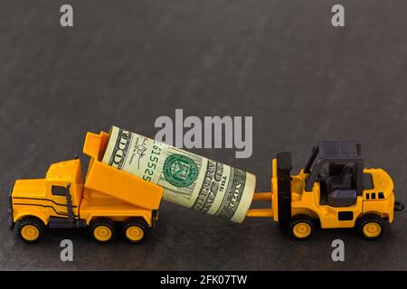 Gelber Muldenkipper mit Dollargeld, der auf schwarzem Steinhintergrund zum Gabelstapler geschickt wird. Geldtransport über Mini-Fahrzeuge. Stockfoto