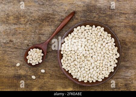 Weiße Erbsenbohnen, auch Navy Bean, Pearl Haricot, Boston Bean in Tonschale auf Holzhintergrund, Draufsicht (Phaseolus vulgaris) Stockfoto