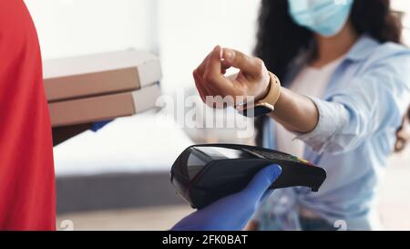 Einfaches Kontaktloses Pay-Konzept. Nahaufnahme des Lieferers in Schutzhandschuhen mit Zahlungsterminal und Pizzakartons, Frau in medizinischer Chirurgie Stockfoto