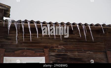 Eiszapfen hängen an einem frostigen, bewölkten Tag vom Dach eines Holzhauses. Das Schieferdach ist gegen den grauen Himmel. Abnormales Wetterkonzept. Neujahr und Weihnachten sind vorbei. Stockfoto