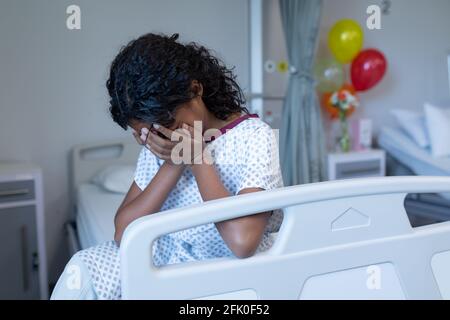 Trauriges, krankes Mädchen mit gemischter Rasse, das weinend auf dem Krankenhausbett sitzt und den Kopf in den Händen hält Stockfoto