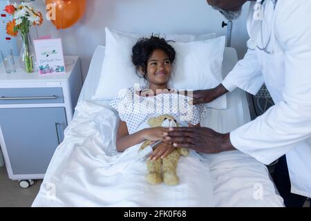 afroamerikanischer männlicher Arzt beruhigend lächelndes krankes Mischling Mädchen Im Krankenhausbett halten Teddy Stockfoto