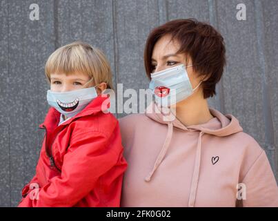 Mutter und Sohn in medizinischen Masken mit gemaltem Lächeln auf ihnen. Eine Frau und ein Kind tragen während der COVID-19-Pandemie Masken. Virenschutz. Positiv dünn Stockfoto