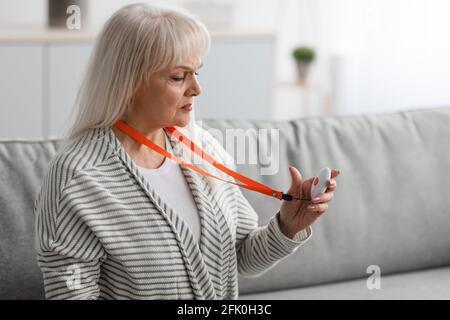 Porträt einer reifen Frau, die einen persönlichen Alarmknopf für einen Notfall auf ihrer Brust hält und zu Hause auf der Couch sitzt. Ältere Dame schaut auf Notsignal eq Stockfoto