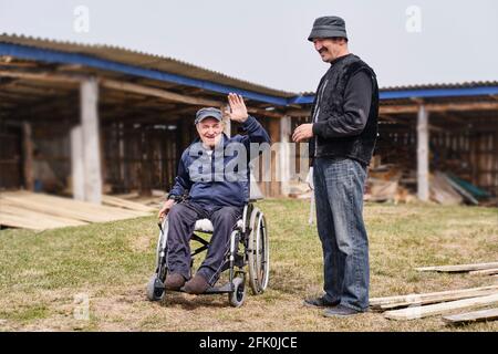 Ein glücklicher älterer Mann in legerer Kleidung in einem Rollstuhl im Hof seines Hauses lächelte und schaute auf die Kamera. Sein Freund steht neben ihm. Stockfoto