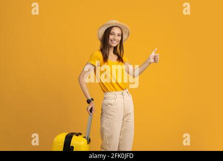Glückliche junge Frau im Sommerhut, die Koffer hält, Daumen zeigt, Platz für Text auf gelbem Studiohintergrund kopieren. Eine hübsche Dame, die uns von Touristen empfohlen wurde Stockfoto