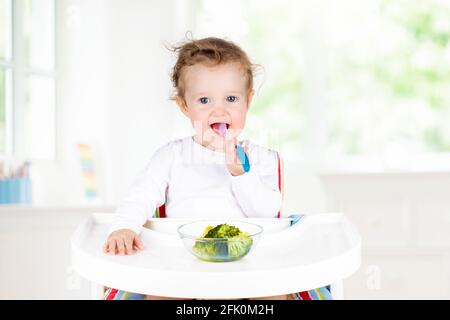 Baby essen Gemüse in der weißen Küche. Kind sitzt auf weißem Hochstuhl. Feste Nahrung für Kleinkinder. Kleines Mädchen essen gesundes Gemüse Mittagessen Stockfoto