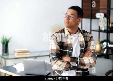 Porträt eines attraktiven erfolgreichen nachdenklichen jungen hispanischen Mannes mit Brille, stilvoll lässig gekleidet, neben dem Schreibtisch stehend mit gekreuzten Armen, rücksichtsvoll zur Seite blickend Stockfoto