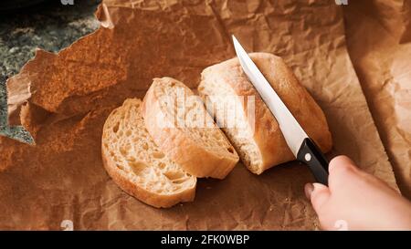 Der Koch schneidet die Ciabatta mit einem Messer in Scheiben. Ciabatta-Scheiben auf Bastelpapier. Frisches, köstliches Gebäck. Frisches, hausgemachtes Brot. Gesunde Ernährung Stockfoto