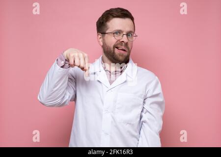 Der überraschte junge Arzt zeigt mit dem Zeigefinger nach unten und sieht mit einer seltsamen Grimasse verwirrt aus. Rosa Hintergrund. Stockfoto