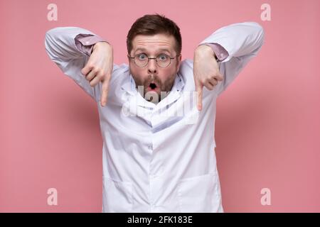 Erschrocken, schockiert, zeigt der Arzt Zeigefinger nach unten, hebt die Hände hoch und sieht mit großen Augen und offenem Mund aus. Rosa Hintergrund. Stockfoto