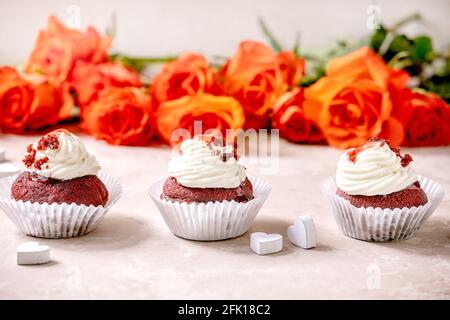 Hausgemachte Cupcakes aus rotem Samt mit Schlagsahne in Reihe, weiße Serviette mit Band, Rosenblumen, Holzherzen auf rosa Textur-Hintergrund. Valentinstag Stockfoto