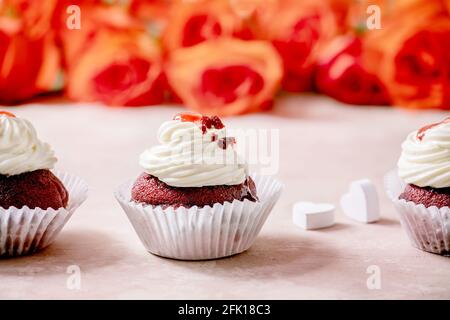 Hausgemachte Cupcakes aus rotem Samt mit Schlagsahne in Reihe, weiße Serviette mit Band, Rosenblumen, Holzherzen auf rosa Textur-Hintergrund. Valentinstag Stockfoto