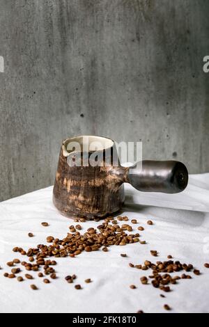 Brown Handwerk Keramik Kaffeekanne türkischen cezve auf weißen Tischdecke mit Kaffeebohnen oben stehen. Graue Wand dahinter. Speicherplatz kopieren. Stockfoto