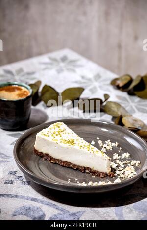 Stück roher veganer Käsekuchen, ohne Backen glutenfrei, verziert mit Limettenschale und Cashewnüssen auf dem Teller. Tasse schwarzen Kaffee und Eukalyptus Zweige auf oder Stockfoto