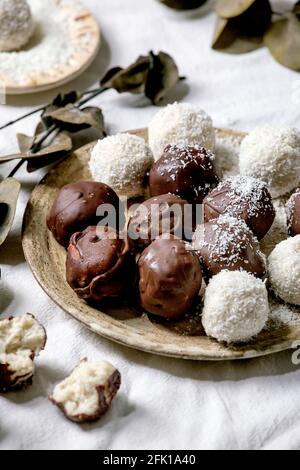 RAW vegan hausgemachte Kokosschokolade Süßigkeiten Kugeln mit Kokosflocken in Keramik-Platte über weißem Textil-Hintergrund mit Eukalyptus-Zweigen. Stockfoto