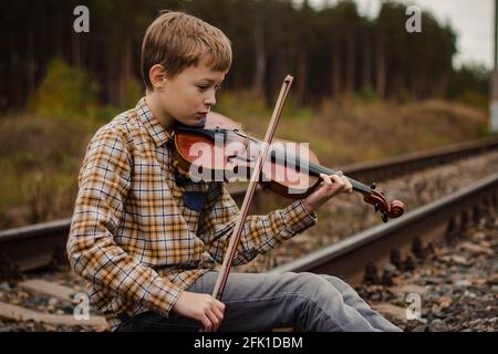 Ein schöner blonder Junge sitzt auf den Schienen der Eisenbahn und spielt Geige. Stockfoto